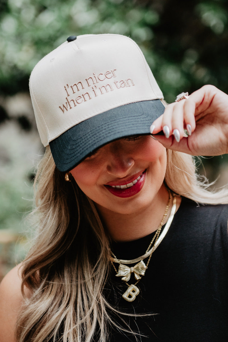Smiling woman in a hat reading "i'm nicer when i'm tan." She wears red lipstick, a black top, layered necklaces, and is outdoors against a blurred green background.