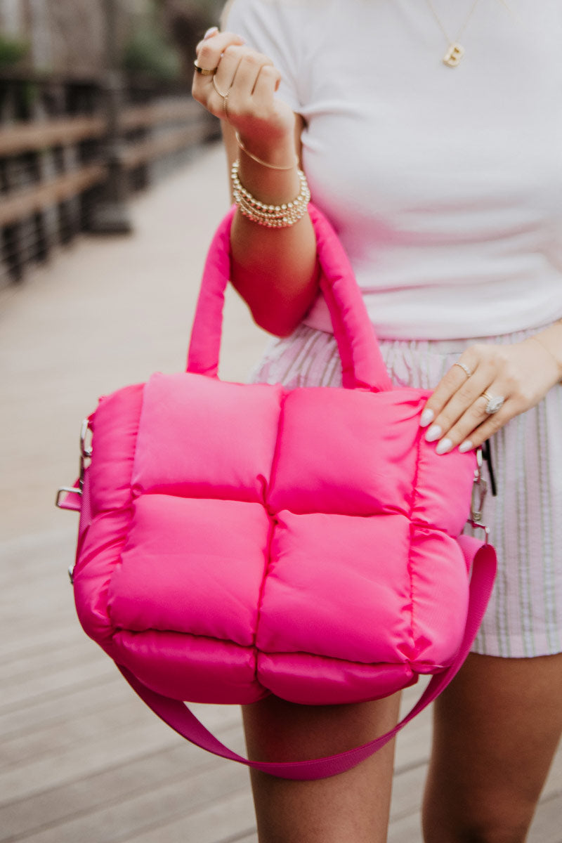 A woman holds a bright pink, quilted handbag with a thick handle and strap. She's wearing a white top and striped shorts.