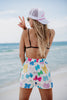 A woman with long blonde hair is standing on a beach, facing the ocean. She wears a white cap, sunglasses, and colorful bow-printed shorts, making a peace sign.
